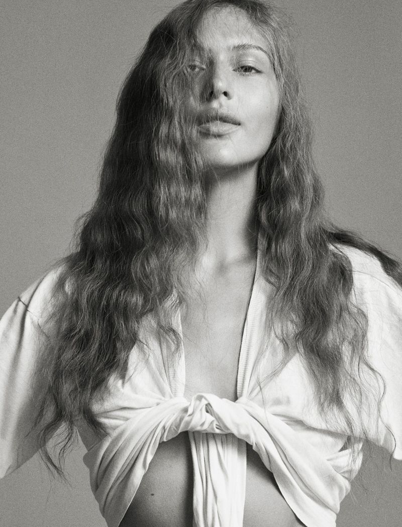Model Angelina Kendall in a tied white t shirt with long wavy hair against a grey seamless background