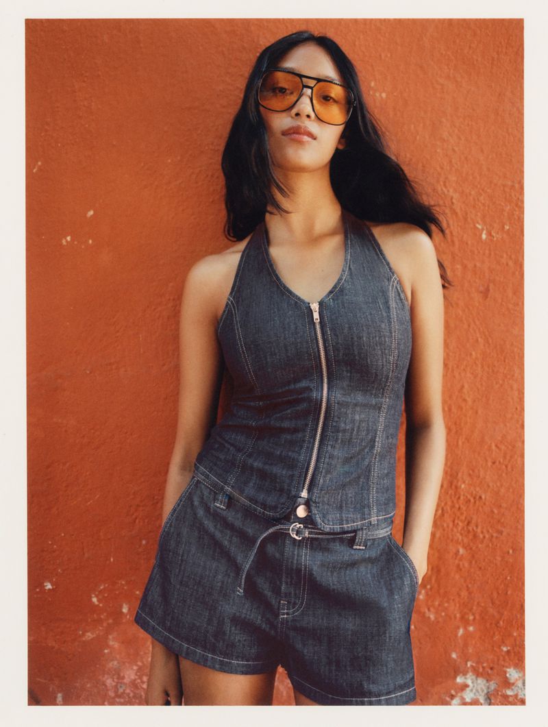 A woman in a denim halter top and shorts poses against an orange wall, accessorized with orange sunglasses, showcasing a fashion-forward look.