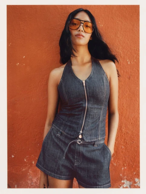 A woman in a denim halter top and shorts poses against an orange wall, accessorized with orange sunglasses, showcasing a fashion-forward look.