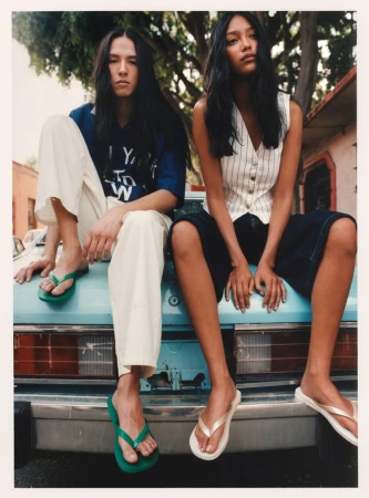 Two models sit on a turquoise car in flip-flops. The long-haired pair wear casual fashions in a sun-drenched outdoor scene with trees.