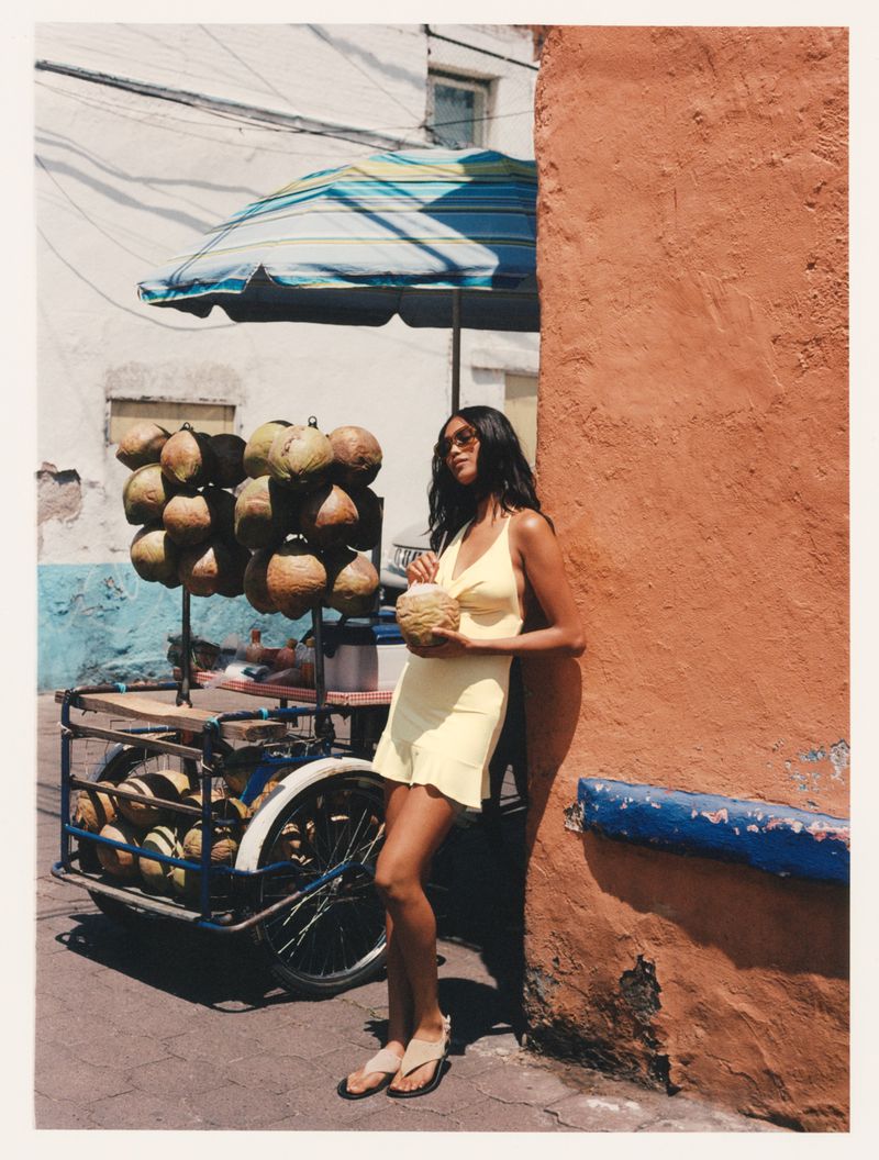 Woman in yellow dress poses with coconut near coconut cart on city street corner, sunny day with shadows.