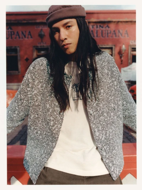 Portrait of a young adult male with long hair wearing a patterned jacket, t-shirt, and hat in an outdoor setting.