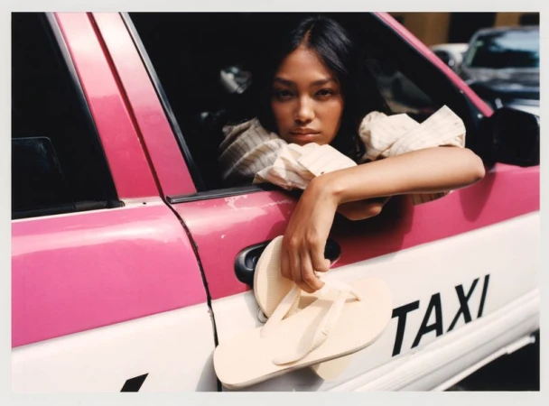 Woman poses inside pink taxi with sandals in hand, soft lighting, eye-level angle. Automotive fashion editorial shot in urban environment.