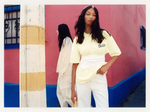 Two Asian women pose in a vibrant urban setting. One wears a yellow top and white pants, the other a white top and pants.
