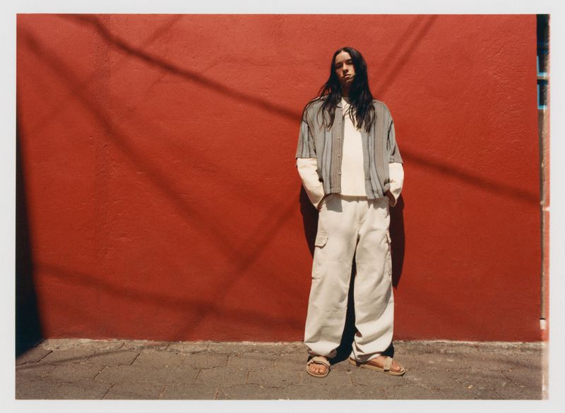 An adult with long, dark hair poses against a red wall in a striped shirt and white pants, creating a casual fashion image.