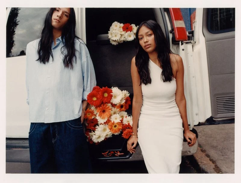 A young man and woman pose next to a white van with a bouquet of colorful gerbera daisies in the foreground, wearing casual clothing.