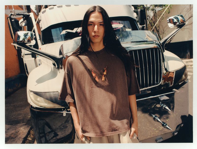 A long-haired person poses in a brown t-shirt in front of a truck with chrome details outdoors on a sunny day.