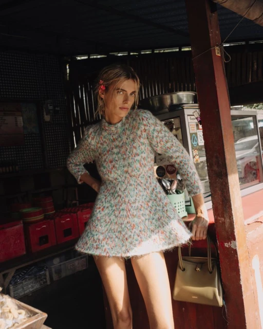Young woman in a short patterned dress poses next to an olive-green handbag in an outdoor, rustic food stand setting with natural light.