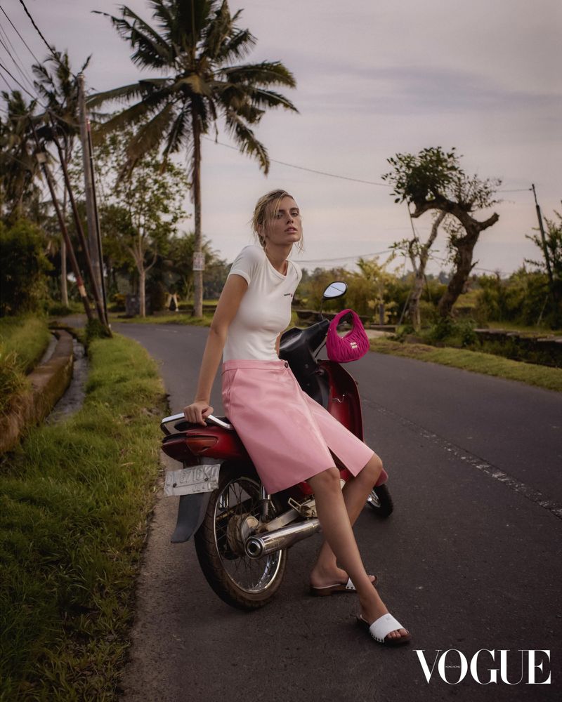 Adult female model sits on a red motorcycle on a rural road lined with grass and palm trees, holding a small pink purse.
