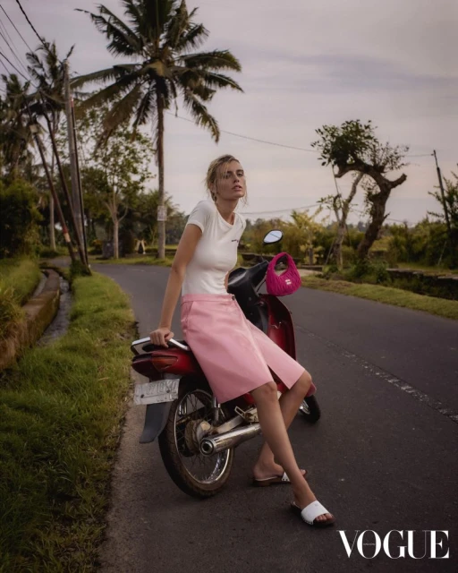 Adult female model sits on a red motorcycle on a rural road lined with grass and palm trees, holding a small pink purse.