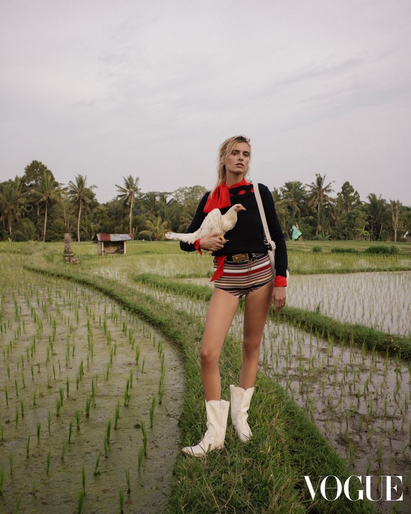 A woman in striped shorts and white boots holds a white chicken in a lush rice field with palm trees, under a cloudy sky, fashion shoot.