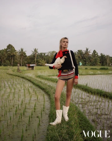 A woman in striped shorts and white boots holds a white chicken in a lush rice field with palm trees, under a cloudy sky, fashion shoot.