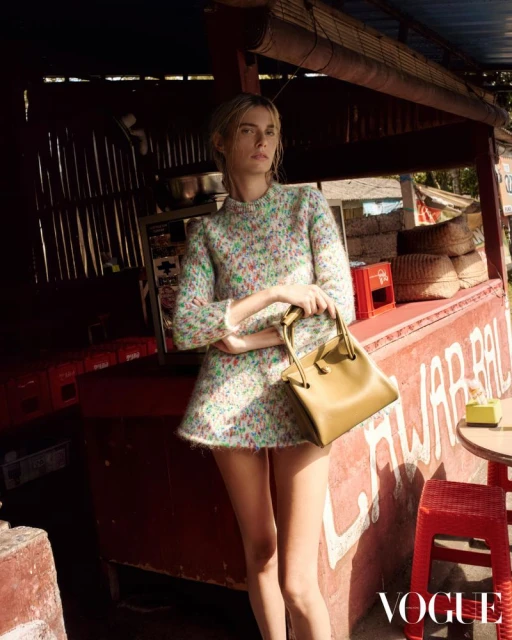 A fair-skinned woman poses in a floral dress with a tan purse, standing in front of an outdoor food stand with 'Vogue' logo in the corner.