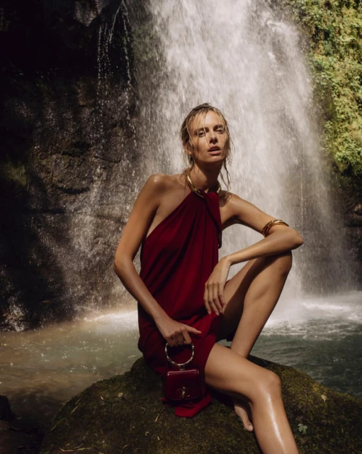 Elegant woman in a red dress poses with a matching purse near a flowing waterfall in a natural setting, complemented by gold jewelry.