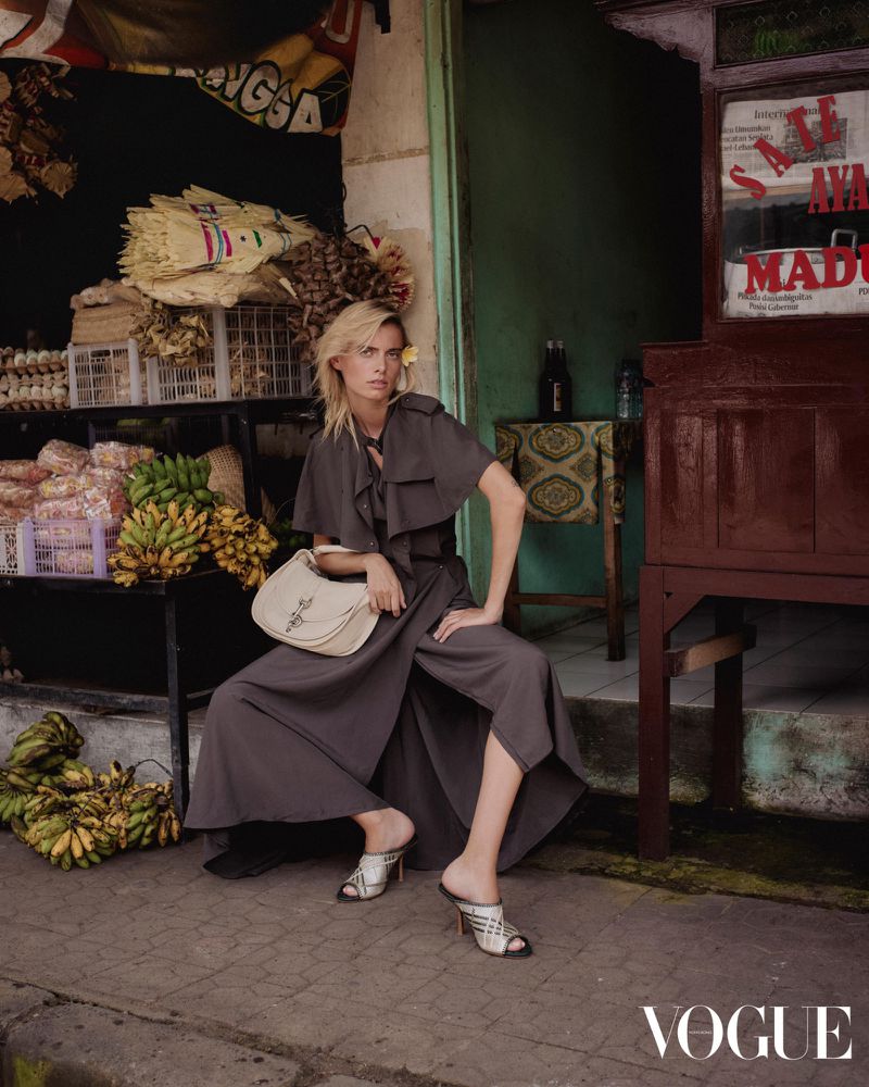 Blonde female fashion model in a gray dress sits in front of a market with bananas and souvenirs, holding a handbag, Vogue logo in the corner.
