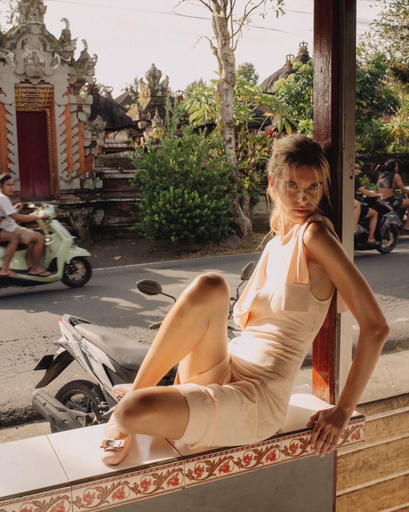 Woman in dress poses on tiled ledge, scooter traffic in background, tropical vibe and outdoor setting with natural light.