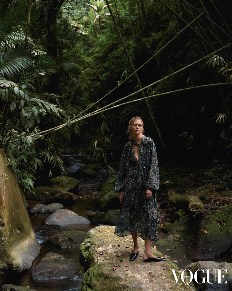 A fashion shot features a blonde woman in a dark floral-print dress standing on rocks near a stream in a lush green jungle setting.