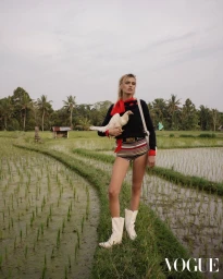 Model wearing a black sweater and striped shorts holding a white bird in a lush green rice paddy field with palm trees under an overcast sky.