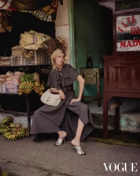 Model wearing a dark grey outfit holding a light handbag while seated near a market stall with bananas