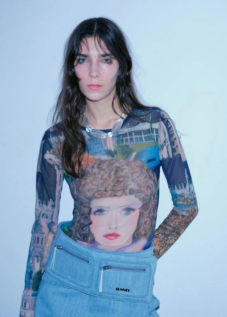 Portrait of a young adult woman in a printed long-sleeve top and denim bag against a plain background in an indoor setting.
