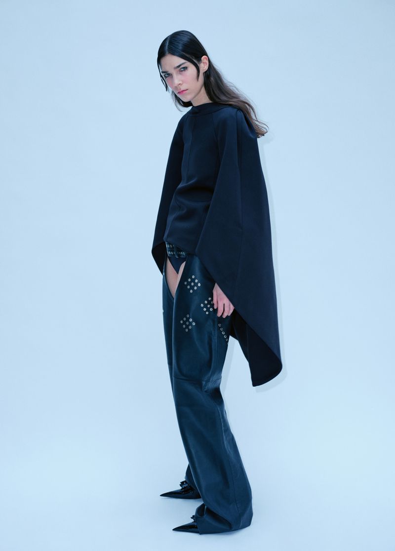Fashion model in a studio wearing a black top and studded leather chaps against a white background in a full shot.