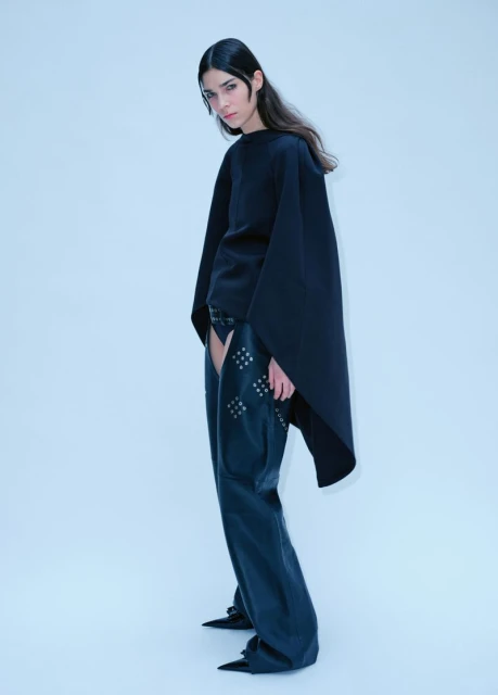 Fashion model in a studio wearing a black top and studded leather chaps against a white background in a full shot.