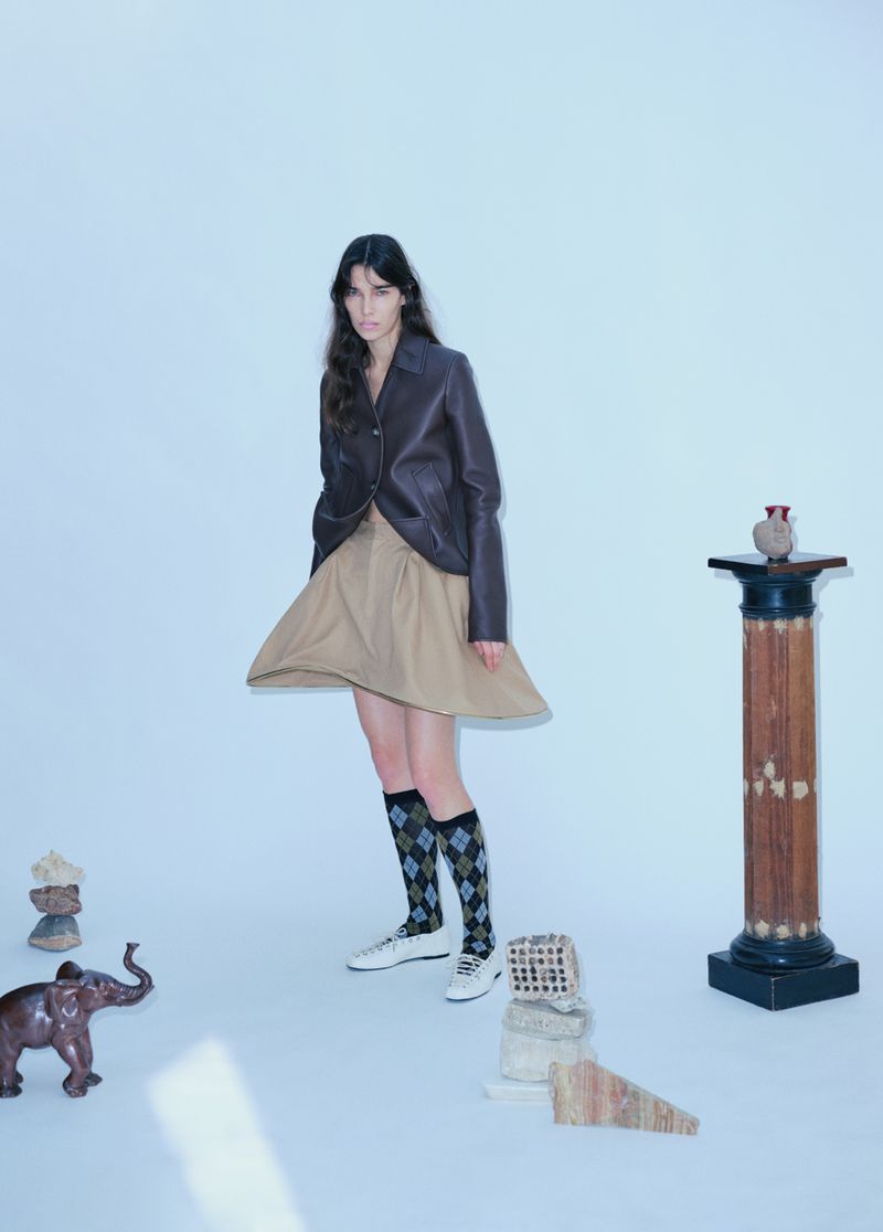 Fashion shot of a young woman wearing a blazer, skirt, and patterned socks, standing in a studio with various decorative objects.