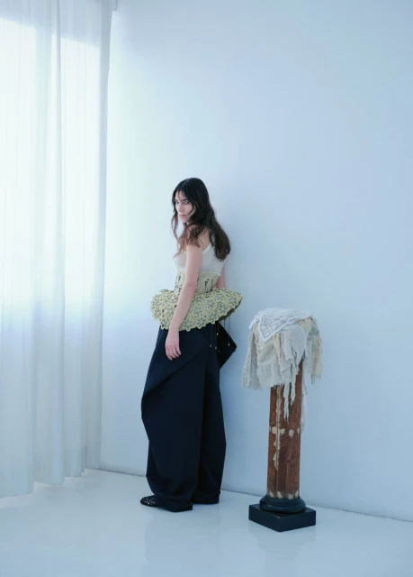 A woman poses in sculptural top and oversized pants against a white wall next to a draped antique column indoors with natural light.