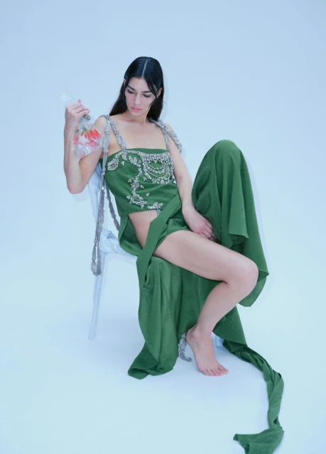A woman sits on a chair in a green dress, holding a fish bowl in a light studio setting with a minimalistic aesthetic.