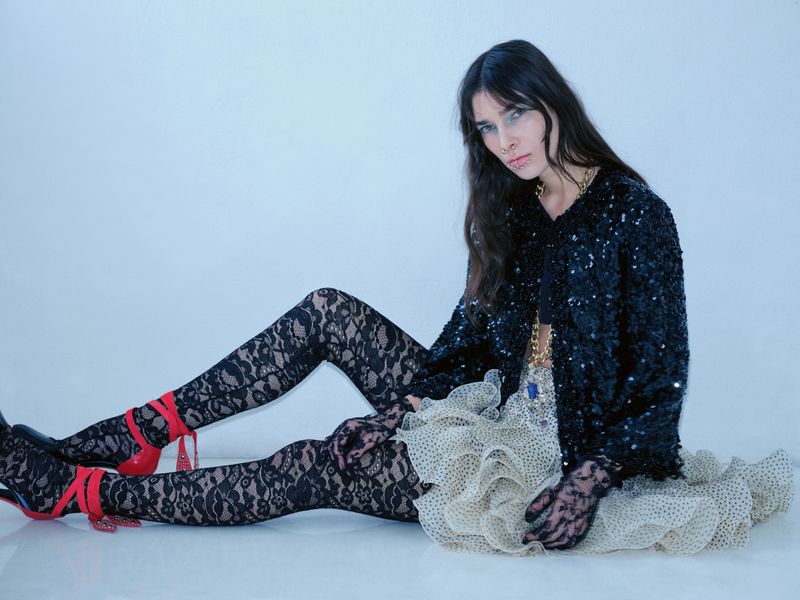 Woman in black sequin jacket and lace tights poses sitting against a white backdrop, wearing red high heels and lace gloves.