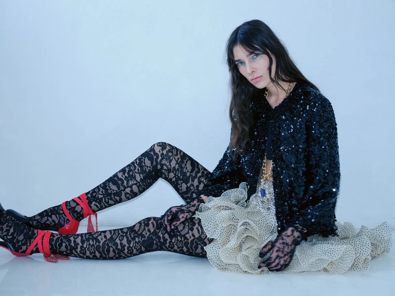 Woman in black sequin jacket and lace tights poses sitting against a white backdrop, wearing red high heels and lace gloves.
