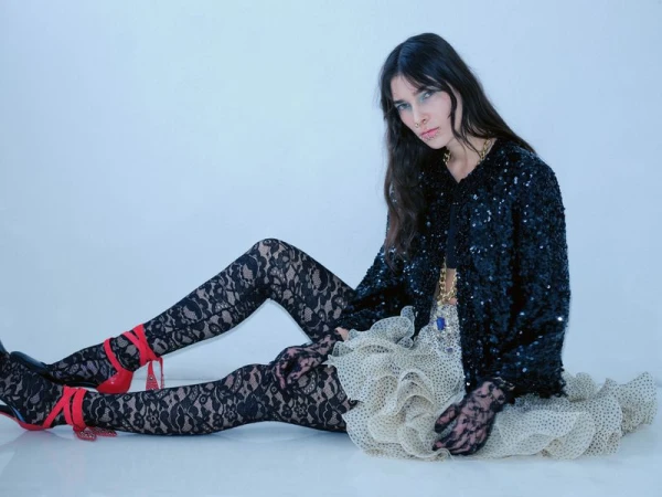 Woman in black sequin jacket and lace tights poses sitting against a white backdrop, wearing red high heels and lace gloves.