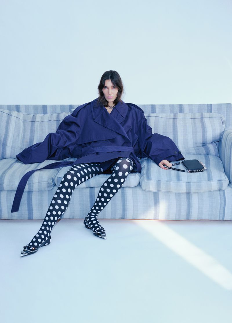 Fashionable woman in blue trench coat and polka dot tights sits on a blue striped couch in a bright indoor studio setting.
