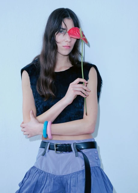 A young woman poses indoors against a light blue wall, holding a red anthurium flower to her face, wearing a black top and lavender skirt.