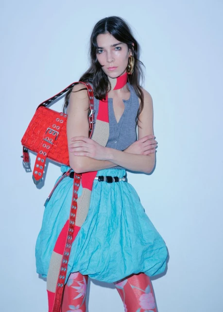 A woman poses in a fashion shot wearing a grey top, blue skirt, tights, and carrying a red purse with a white studio background.