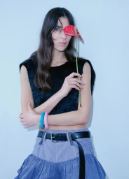 Model holding a red flower covering one eye, wearing a black top and blue skirt against a white studio background.
