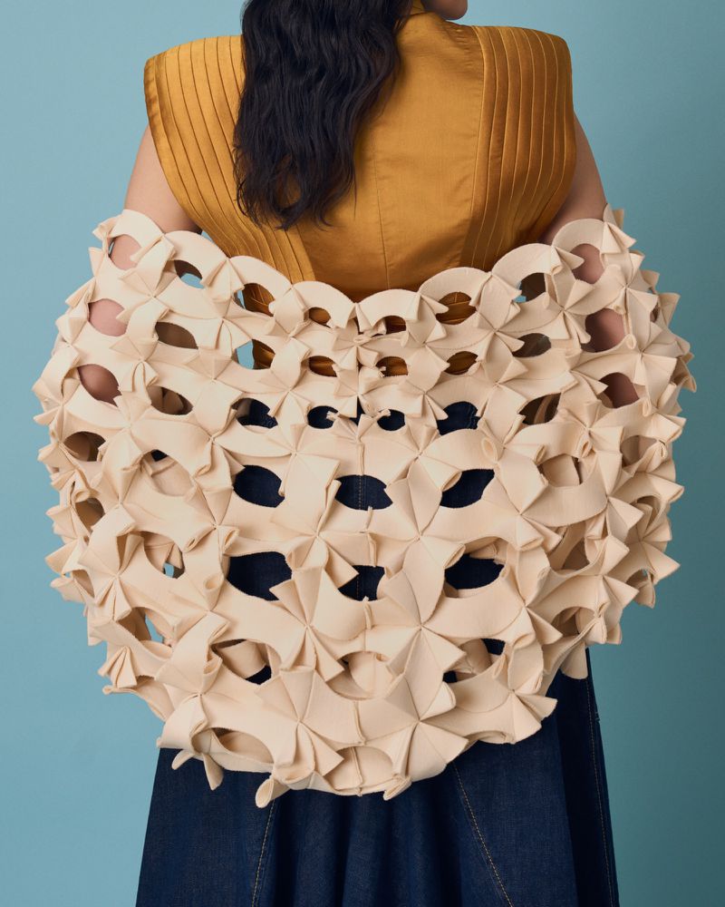 Woman wearing a yellow sleeveless top and an abstract tan jacket with circular cut-outs against a light blue background in a studio shot.