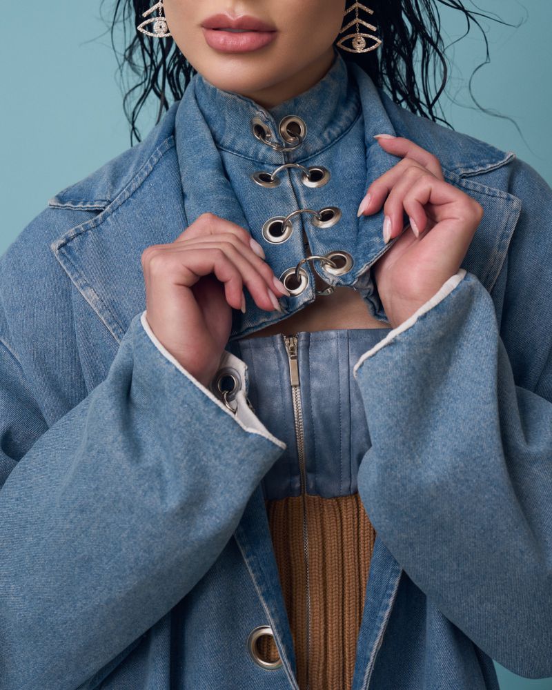 A stylish woman wears a light blue denim jacket and ornate eye-shaped earrings against a light blue background in a studio setting.
