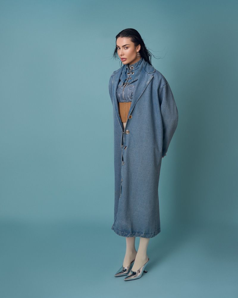 Full-length shot of an adult woman in a blue denim coat, neutral top, white tights, and silver mules against a light blue backdrop.