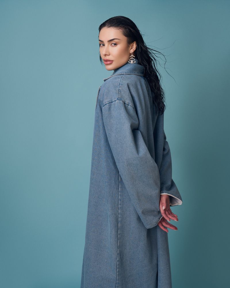 Attractive woman with long dark hair, looking over her shoulder, wearing a denim jacket dress and dangling earrings against a teal backdrop.