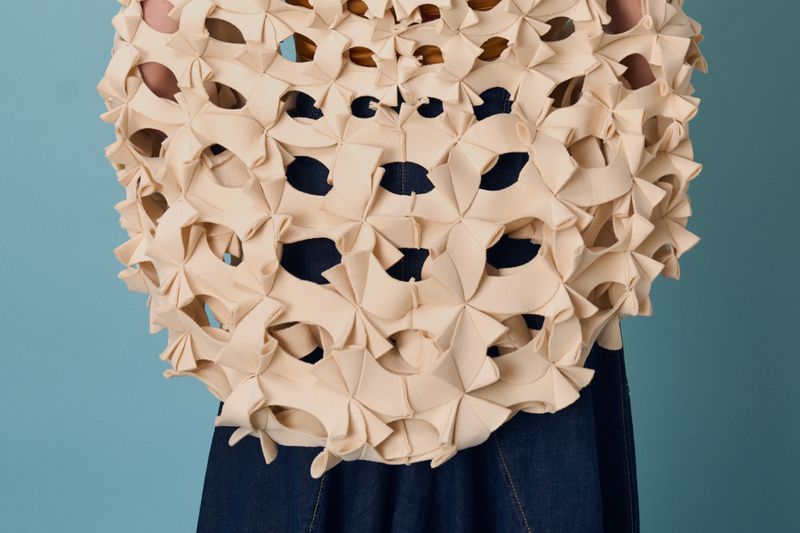 Person wearing a beige, geometric laser-cut top with large holes, paired with high-waisted, indigo-dyed denim pants against a blue backdrop.