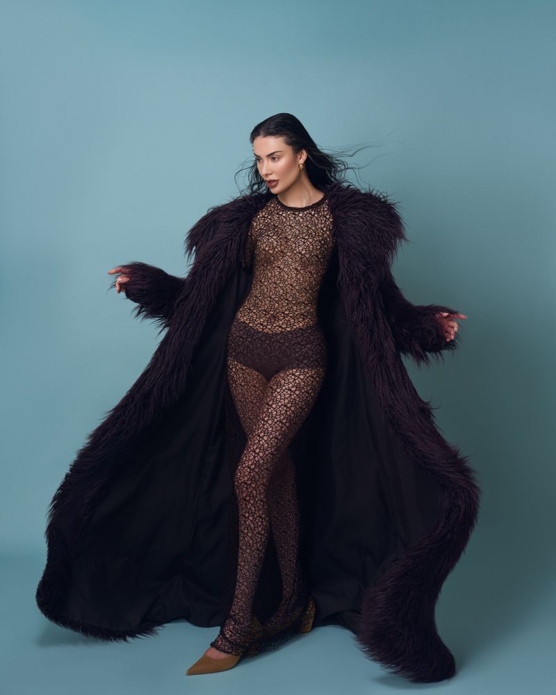Fashionable woman in a long faux fur coat and patterned bodysuit poses against a blue backdrop in a studio setting.