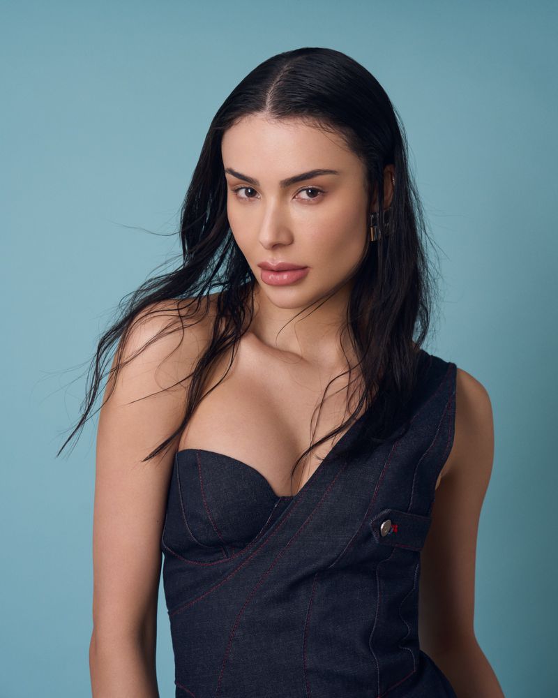 Portrait of a young woman with long black hair wearing a dark blue, asymmetrical denim top against a light blue background.