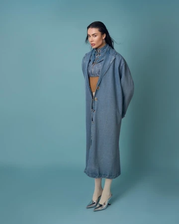 Model wearing a long blue denim coat and metallic silver heels against a solid teal studio backdrop.