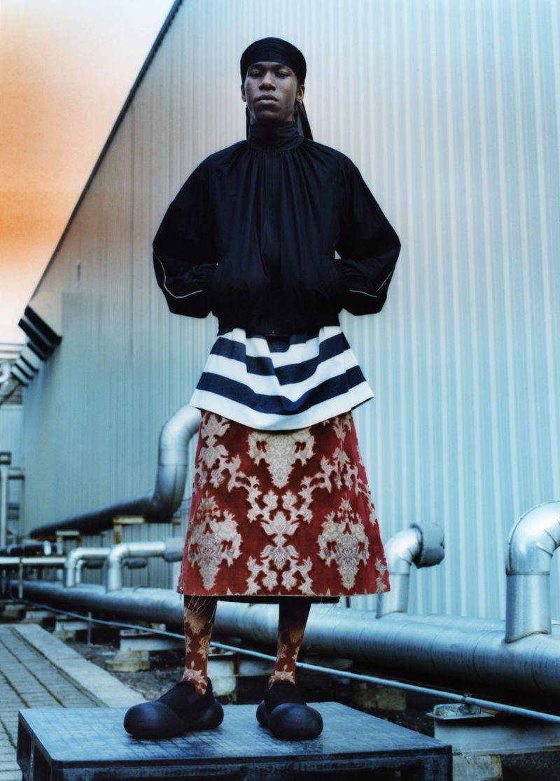Model wearing a black top, striped bib, and patterned skirt stands on a platform with industrial pipes and a warehouse-style building in the background.