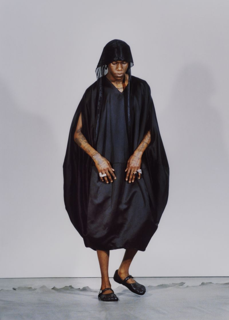 Fashion shot featuring an adult male model in an oversized black dress and head covering, posing in a photo studio with a grey background.