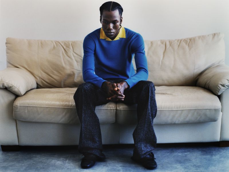 An African American man in a blue sweater and grey patterned pants sits on a cream-colored couch against a light grey background.
