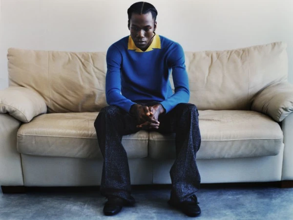 An African American man in a blue sweater and grey patterned pants sits on a cream-colored couch against a light grey background.