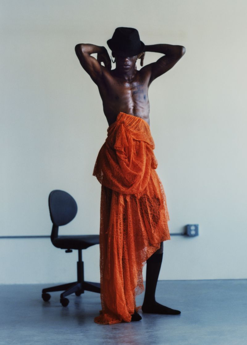 Adult male in studio wearing hat and orange fabric. Dark skin, shirtless, minimal background. Fashion, portrait, studio photography.