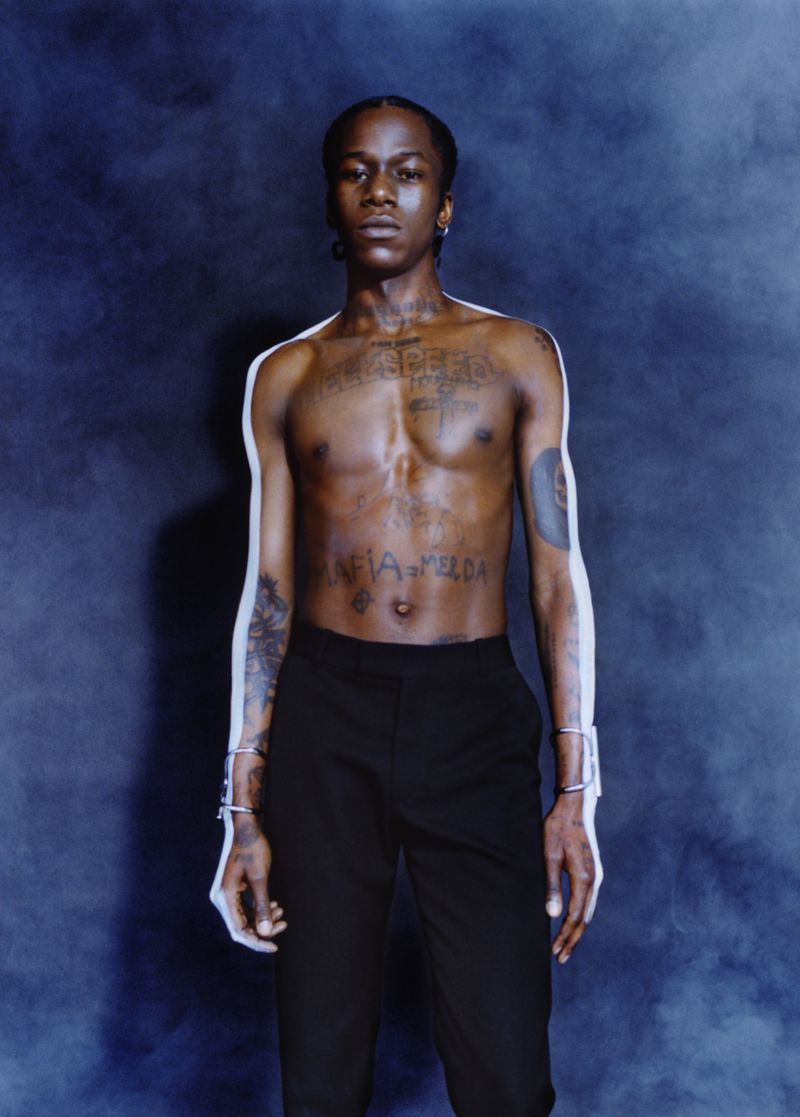 Portrait of a Black adult male with tattoos, wearing pants and bracelets, standing in a dimly lit studio with a smoky background.